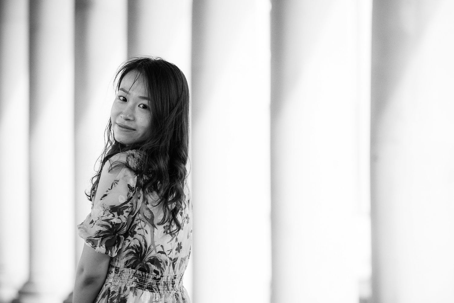 Black and white portrait of a young Asian woman in a floral dress between white columns.