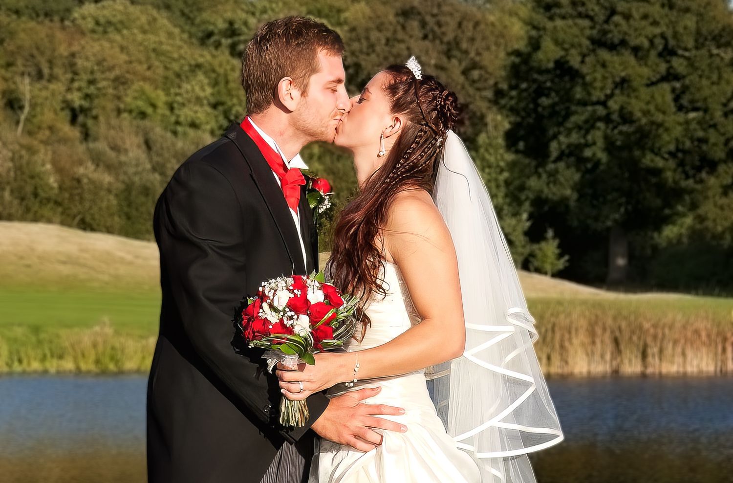 Bride and groom kissing by a lake, bride holding a red rose bouquet in a white wedding dress.
