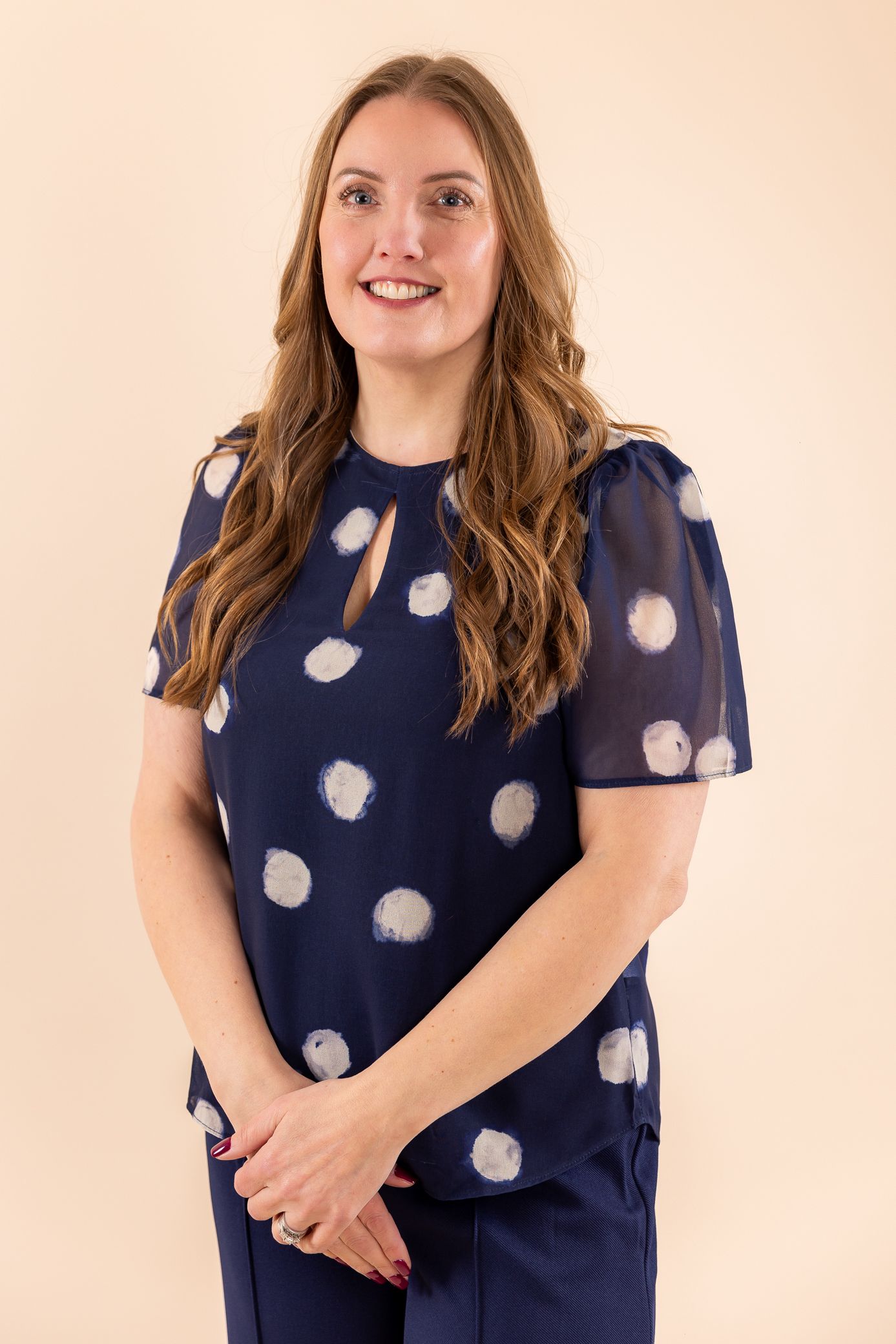 Woman in navy blue polka dot blouse smiling against a beige background.