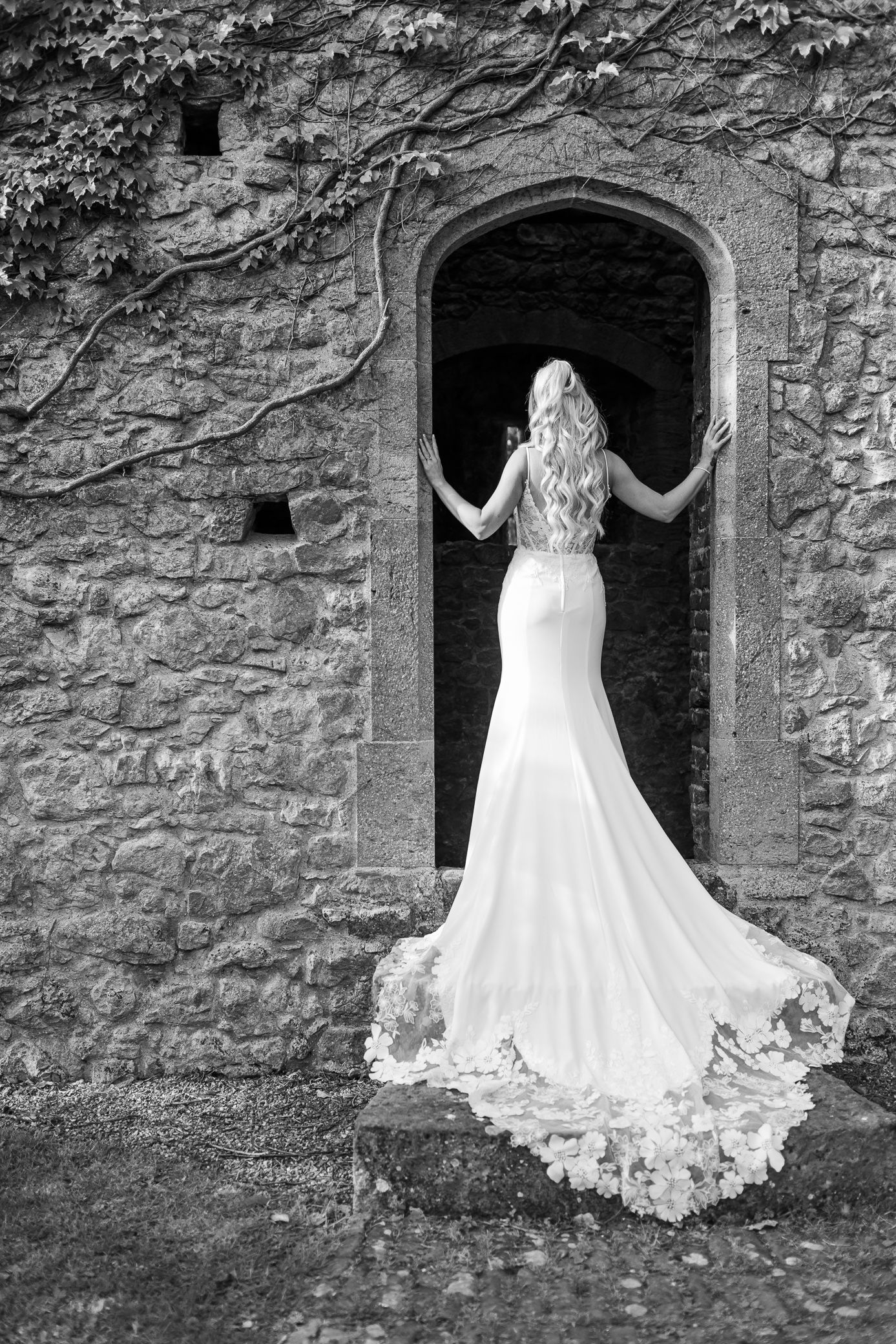 Black and white rear view of bride in lace gown with train at ivy-covered stone arch.