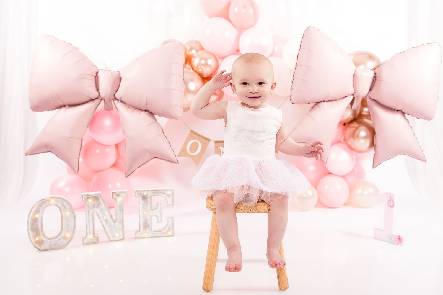 Baby girl in white tutu sits on stool surrounded by pink bow balloons and ONE sign at first birthday party.