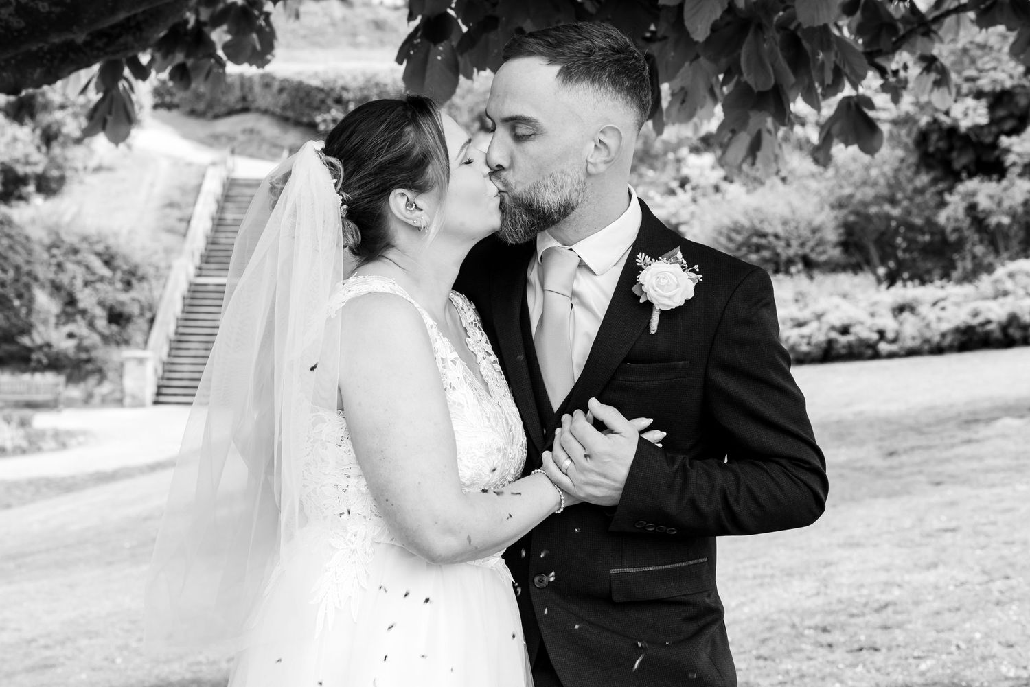 Black and white wedding photo of bride and groom sharing a kiss outdoors under a tree.