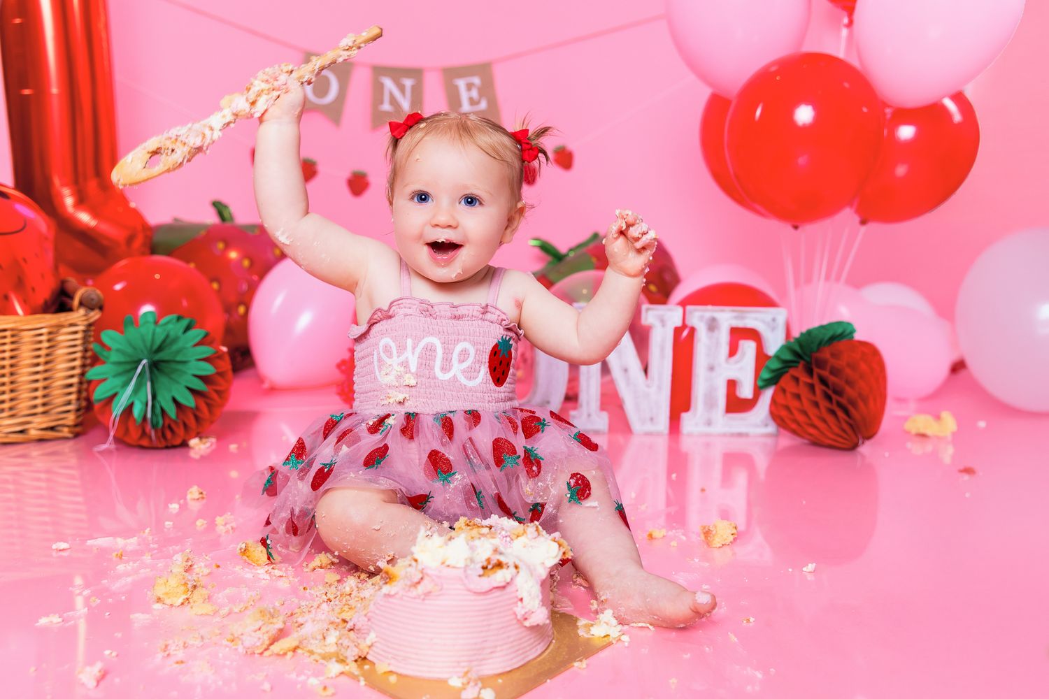 Baby girl in strawberry outfit smashing first birthday cake surrounded by red balloons and ONE decorations on pink backdrop.
