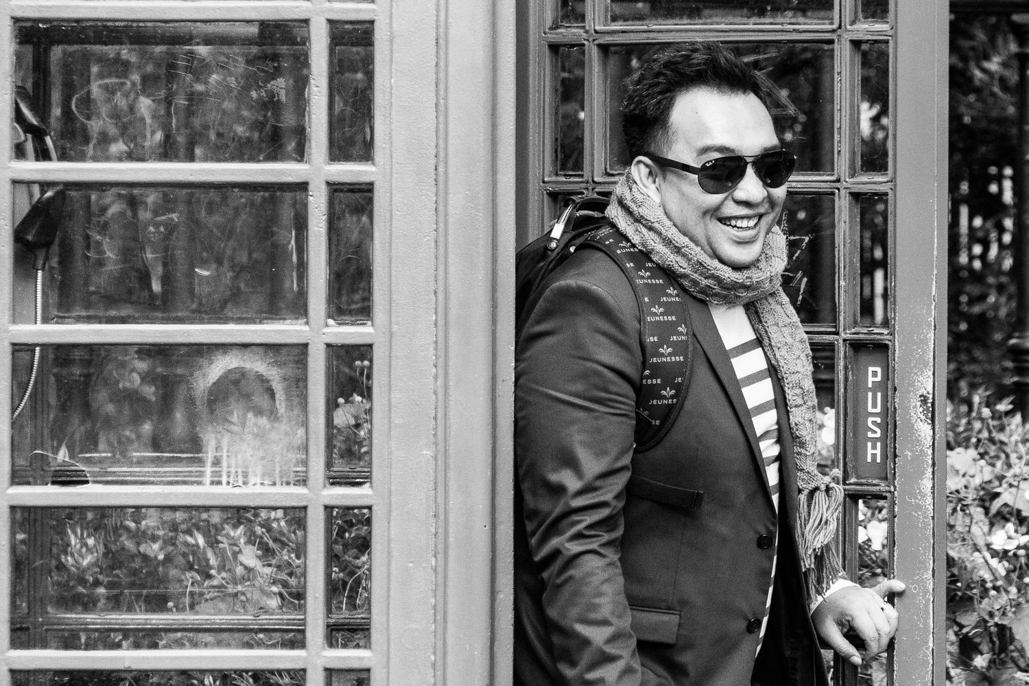 Man in sunglasses and scarf smiling outside a phone booth in black and white street photography.