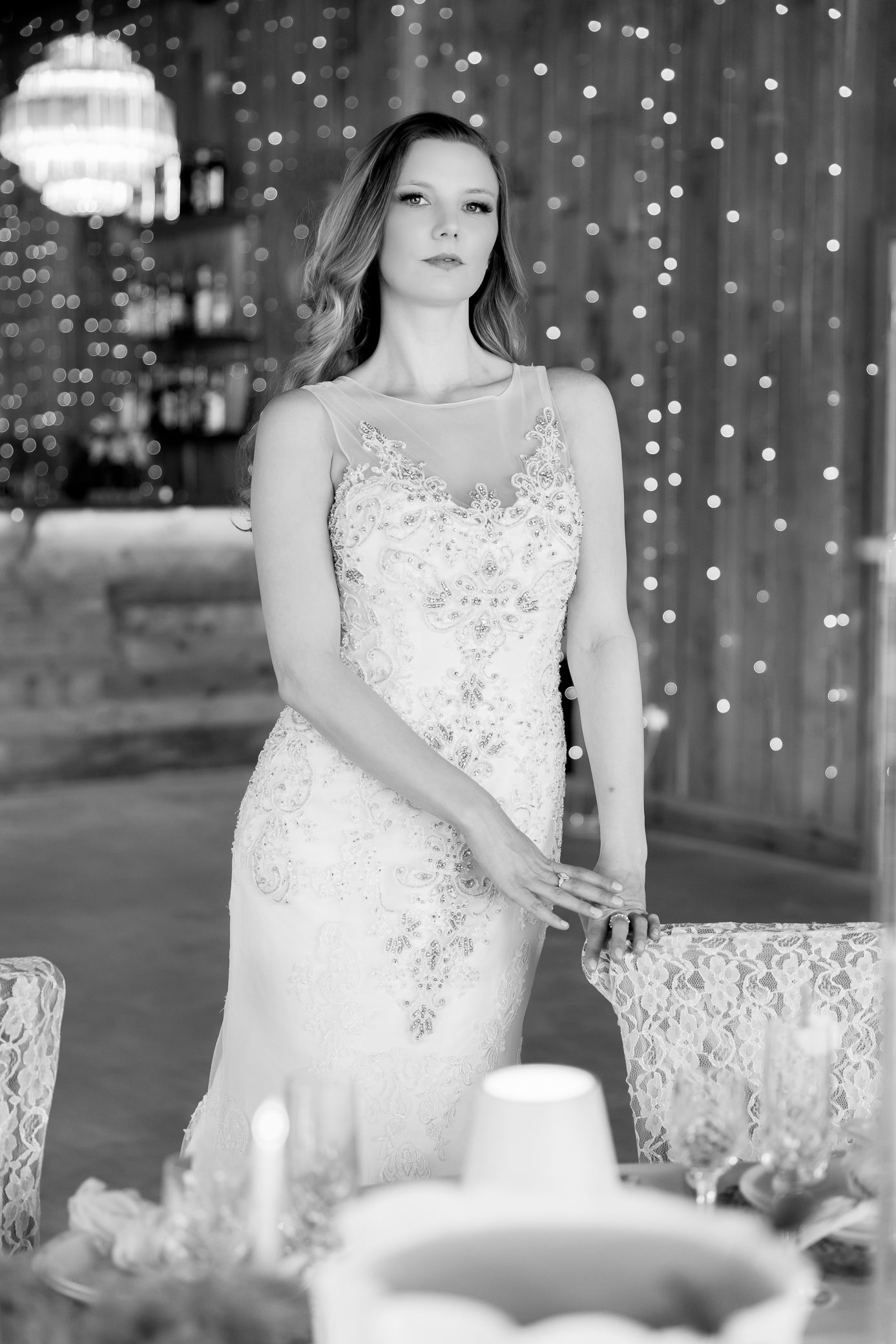 Bride in beaded sheath wedding dress posing by decorated table with fairy lights backdrop.