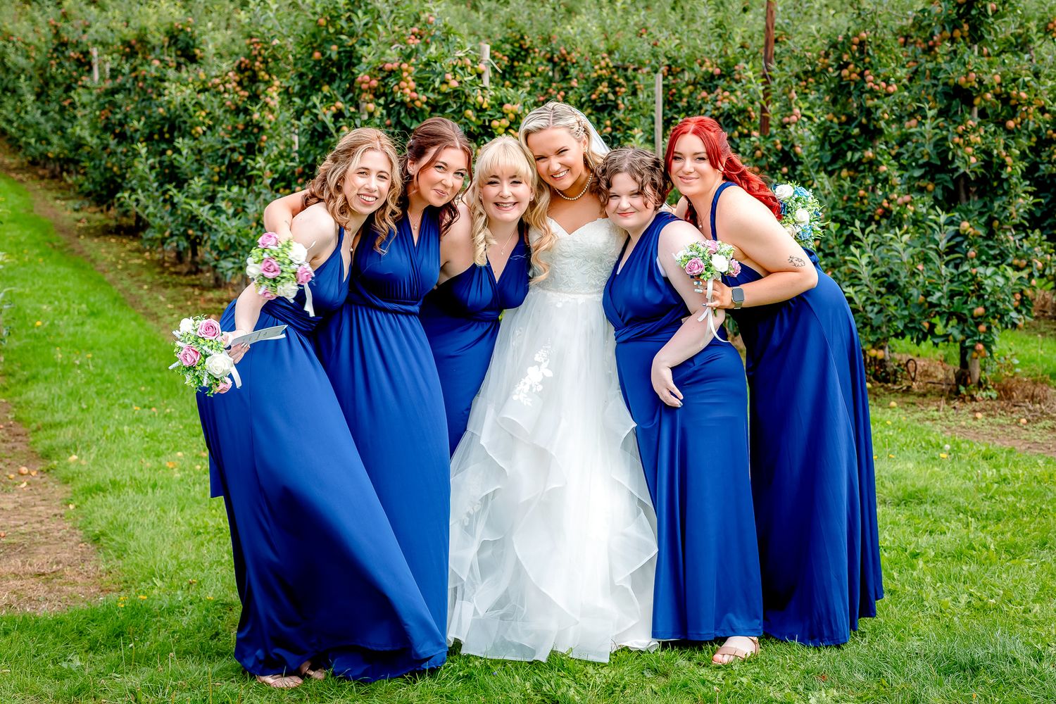 Bride surrounded by five bridesmaids in royal blue dresses posing in an orchard garden setting.