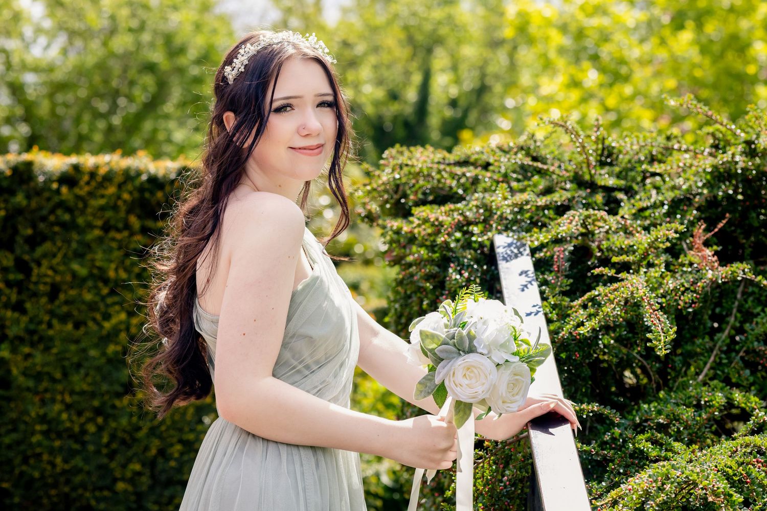 Young woman in sage green bridesmaid dress holding white bouquet, posing outdoors near green hedges.