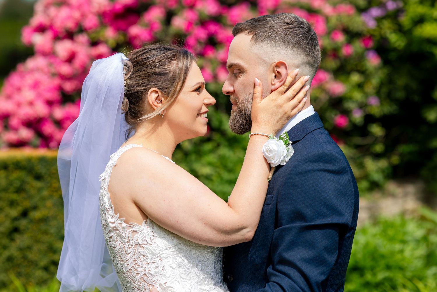 A bride and groom share a romantic moment in front of pink flowers, the bride in a white lace gown and veil.