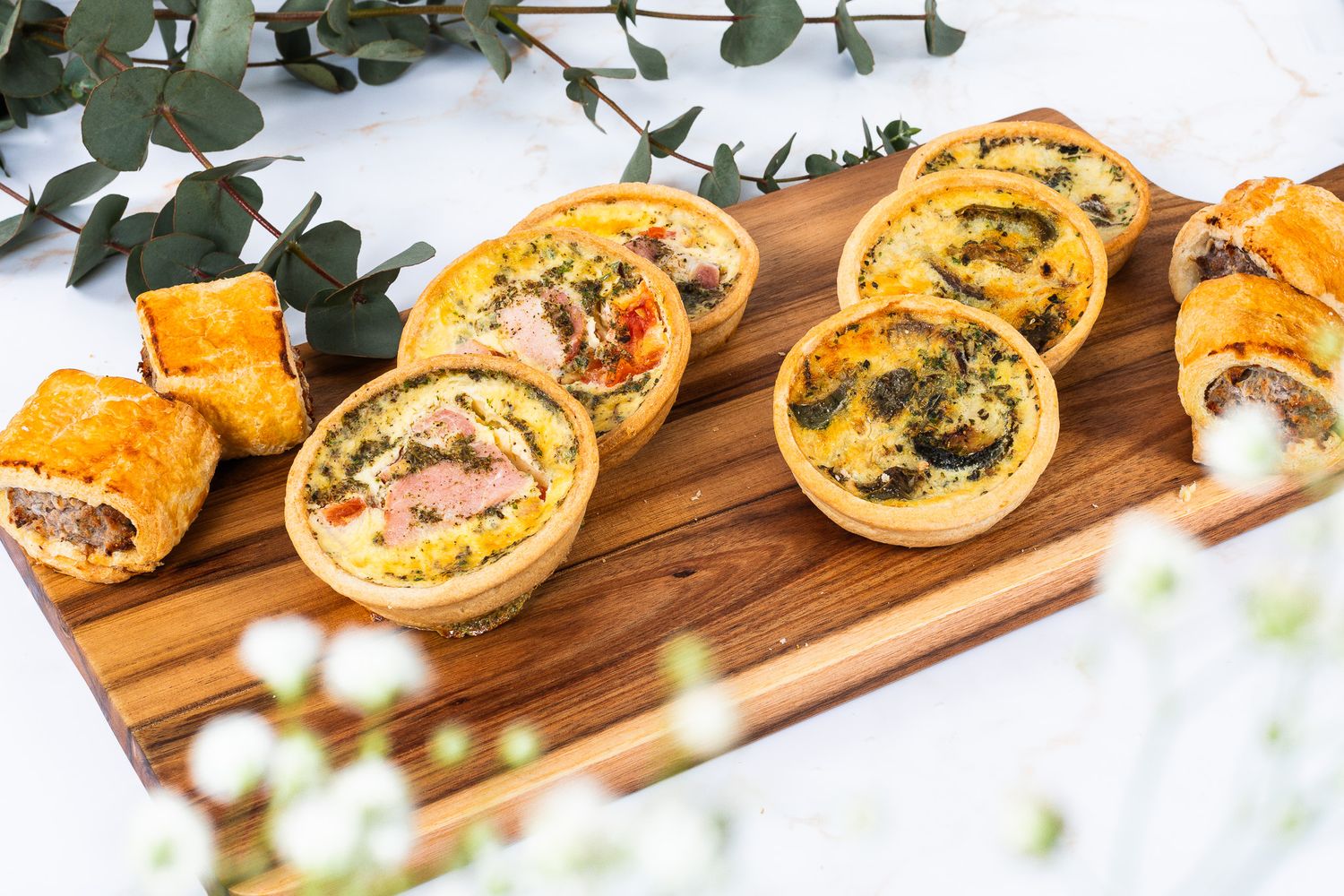 Assorted mini quiches and sausage rolls arranged on a rustic wooden serving board.