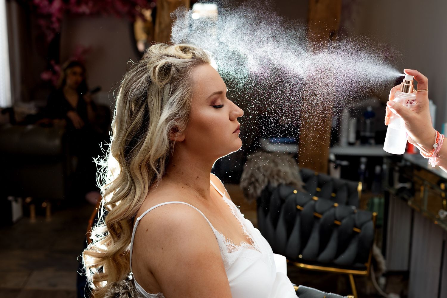 Makeup artist applies setting spray to a blonde bride with curled hair during wedding preparations.