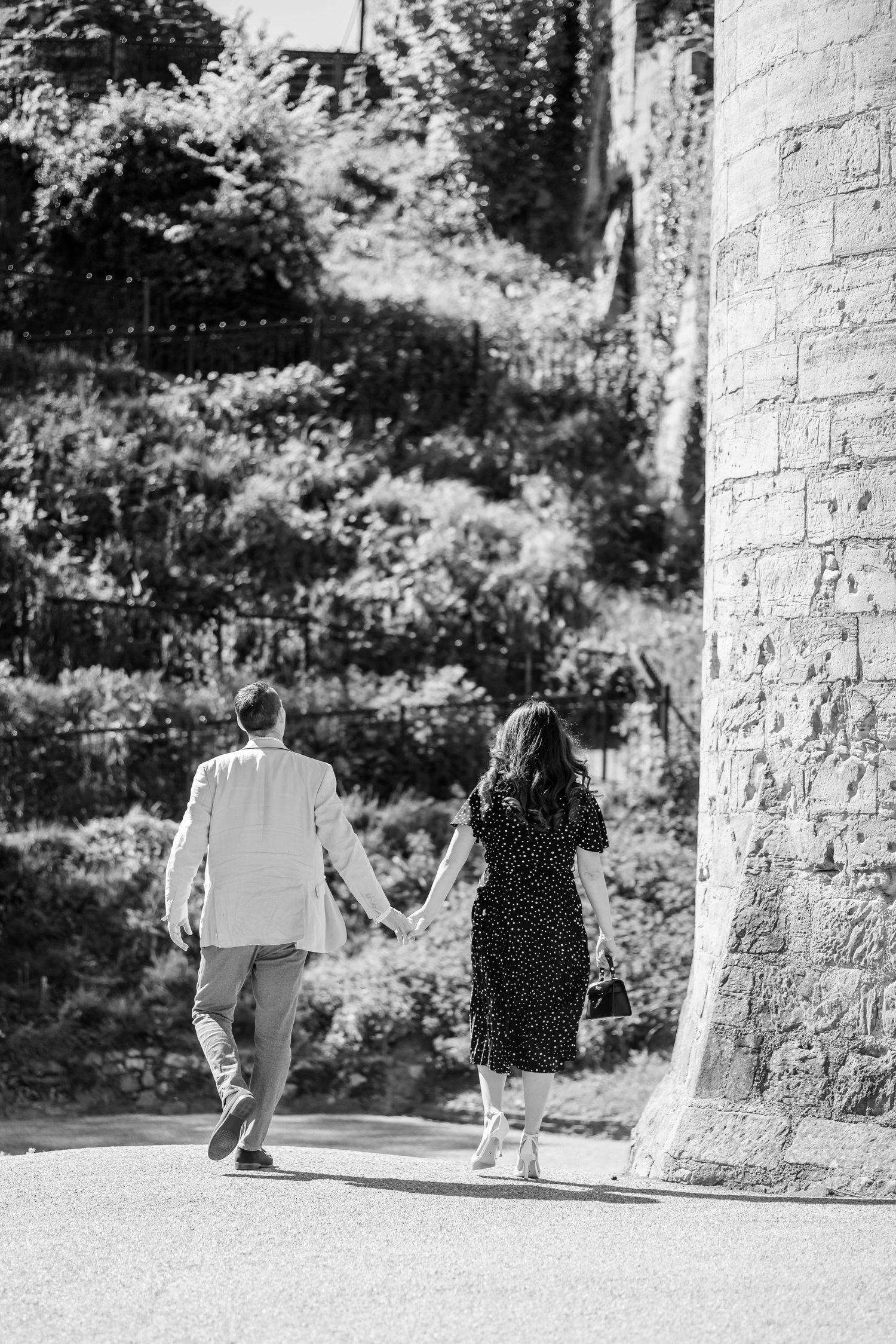 Black and white photo of a couple walking hand in hand past a stone building surrounded by lush greenery.