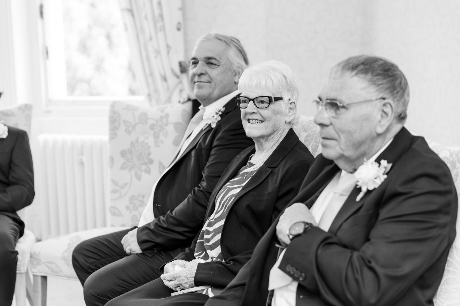 Black and white photo of three elderly wedding guests seated together, smiling during a ceremony.