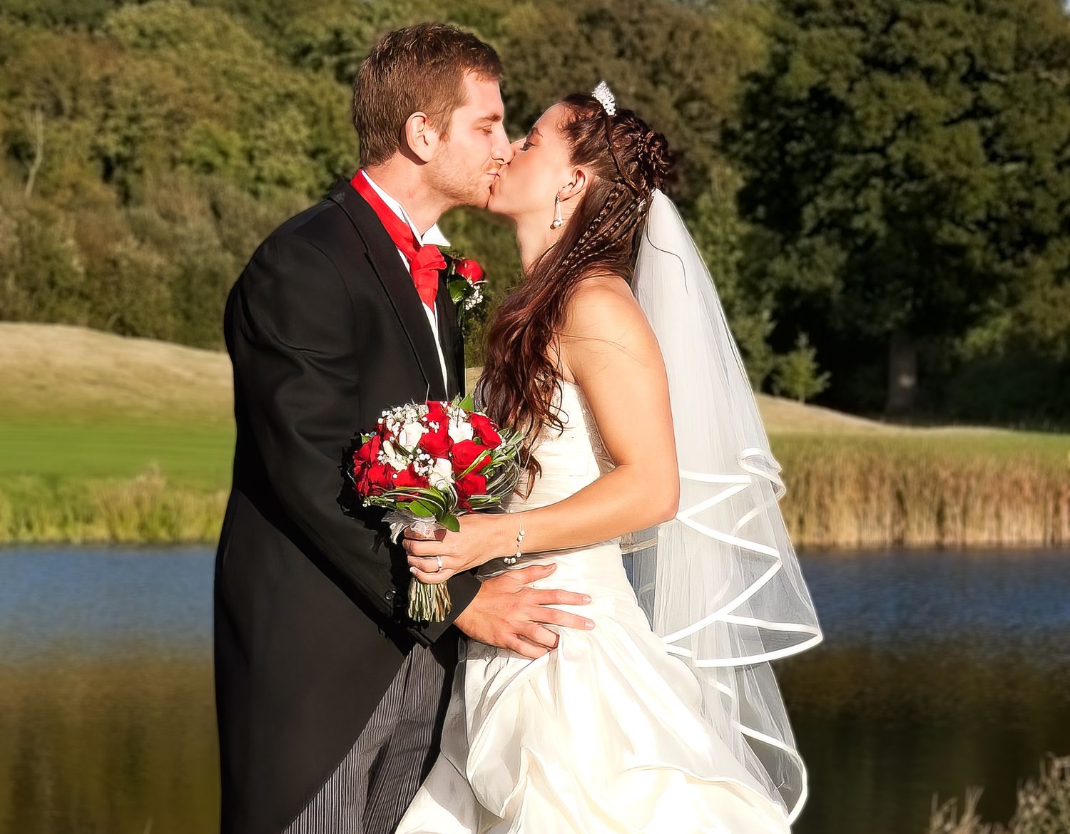 Bride and groom sharing a romantic kiss outdoors, bride holding red rose bouquet by a lake.