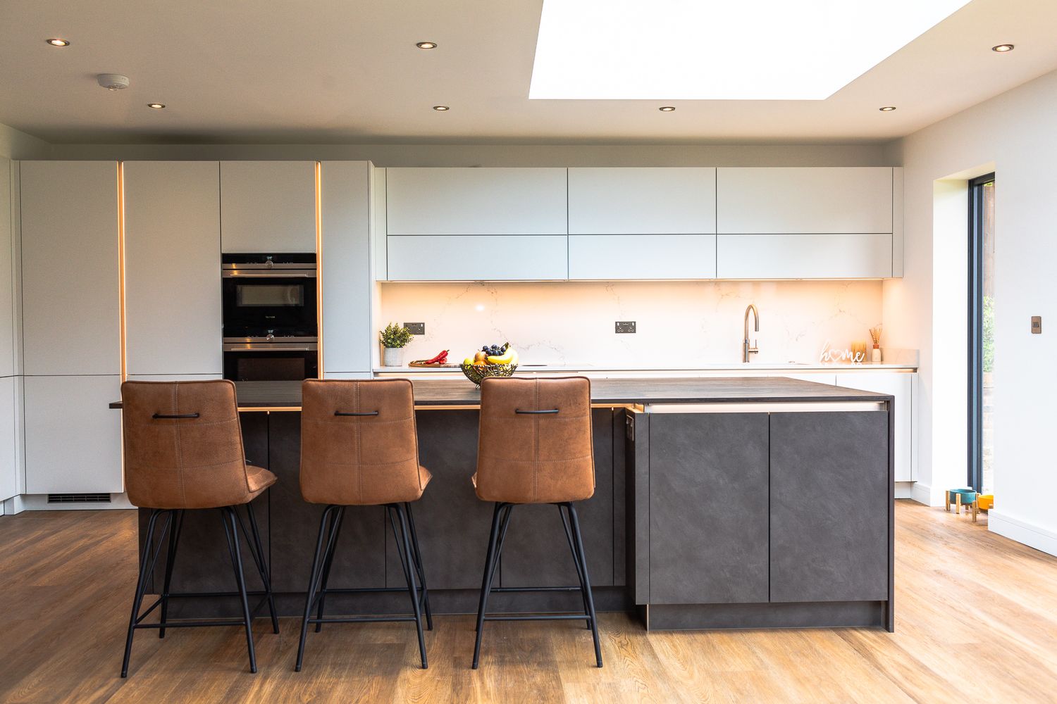 Modern kitchen with grey island, leather bar stools, white cabinets, and skylight overhead.