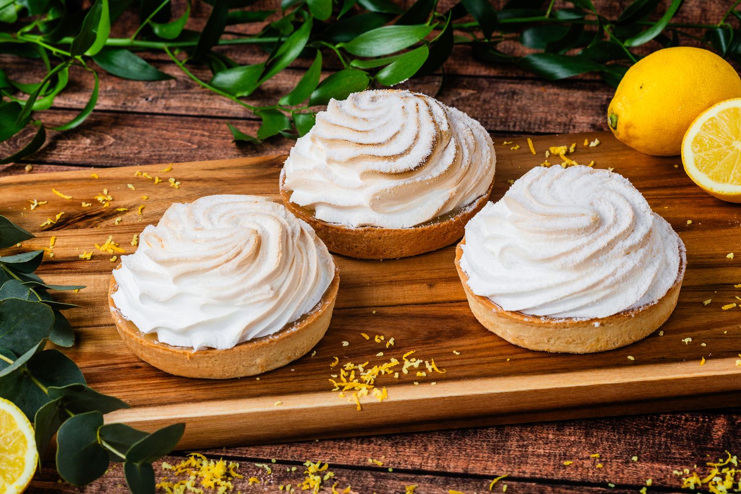 Three lemon meringue tarts with swirled topping on a rustic wooden board with lemons.