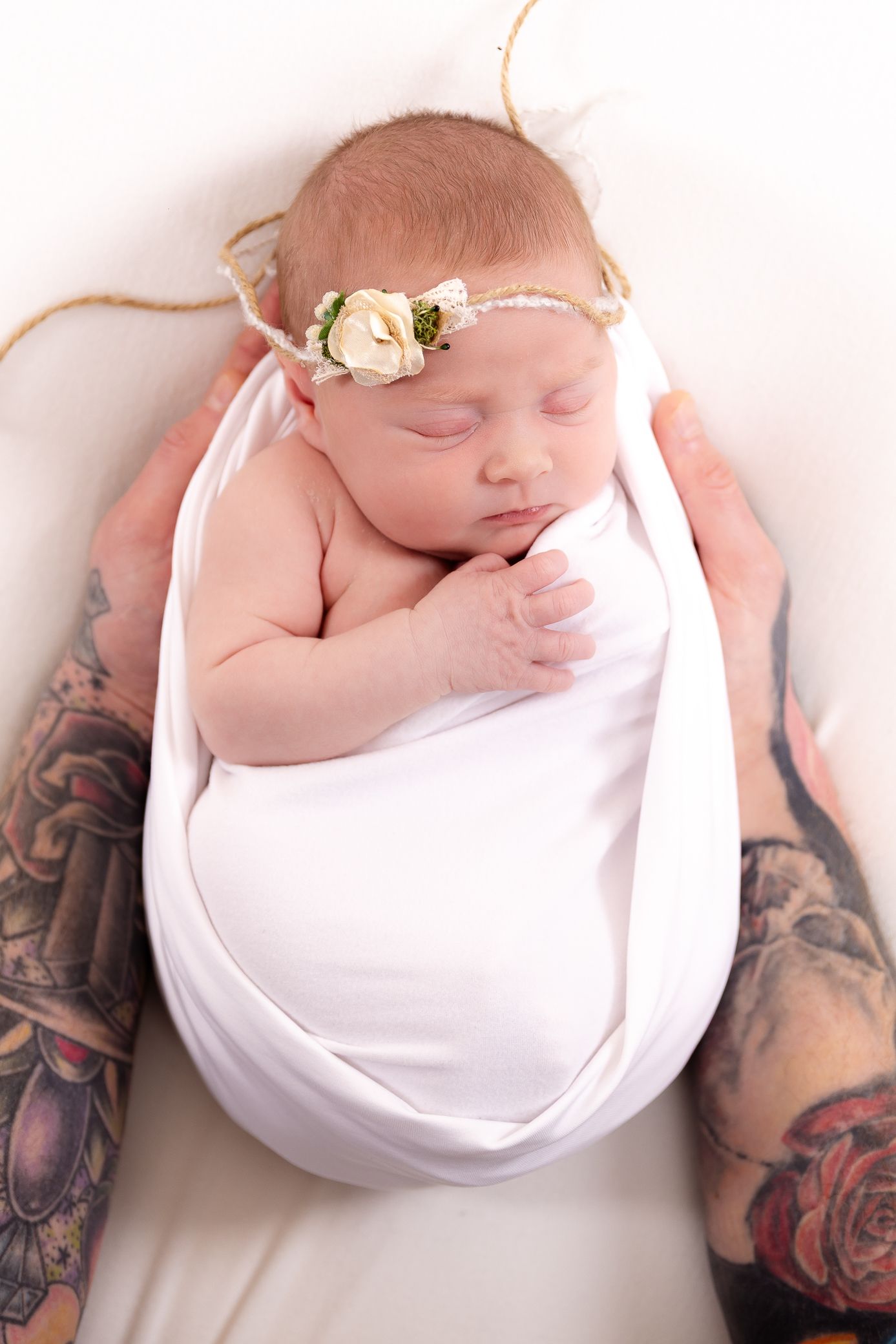 Sleeping newborn baby girl in white swaddle with floral headband cradled by tattooed parents hands.