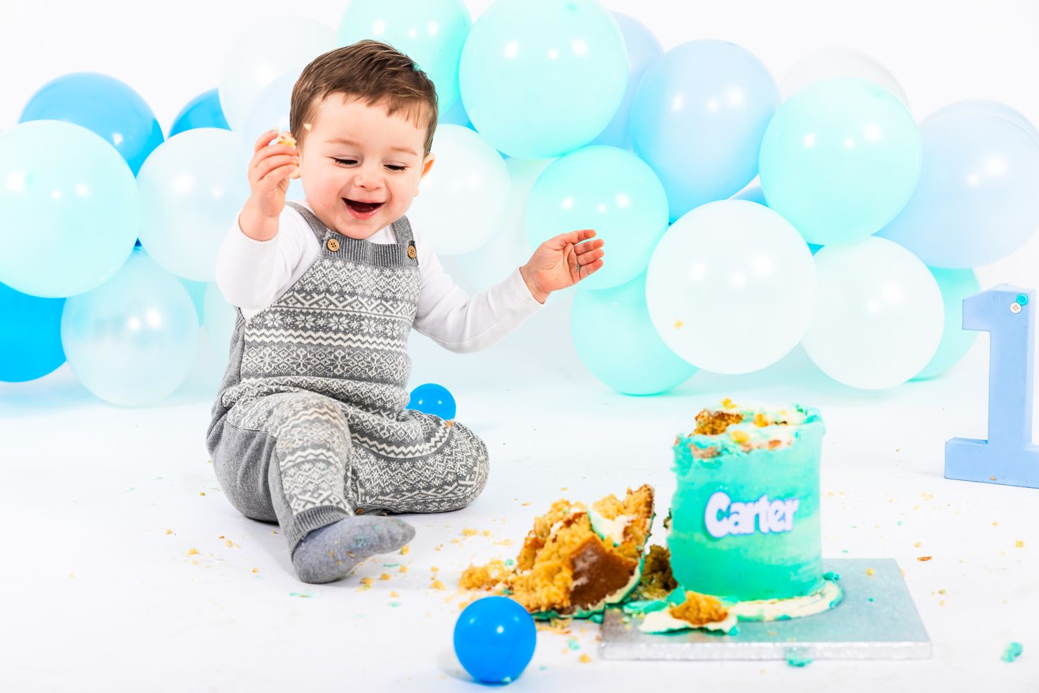 Baby boy named Carter laughing during first birthday cake smash with blue balloons and teal cake.