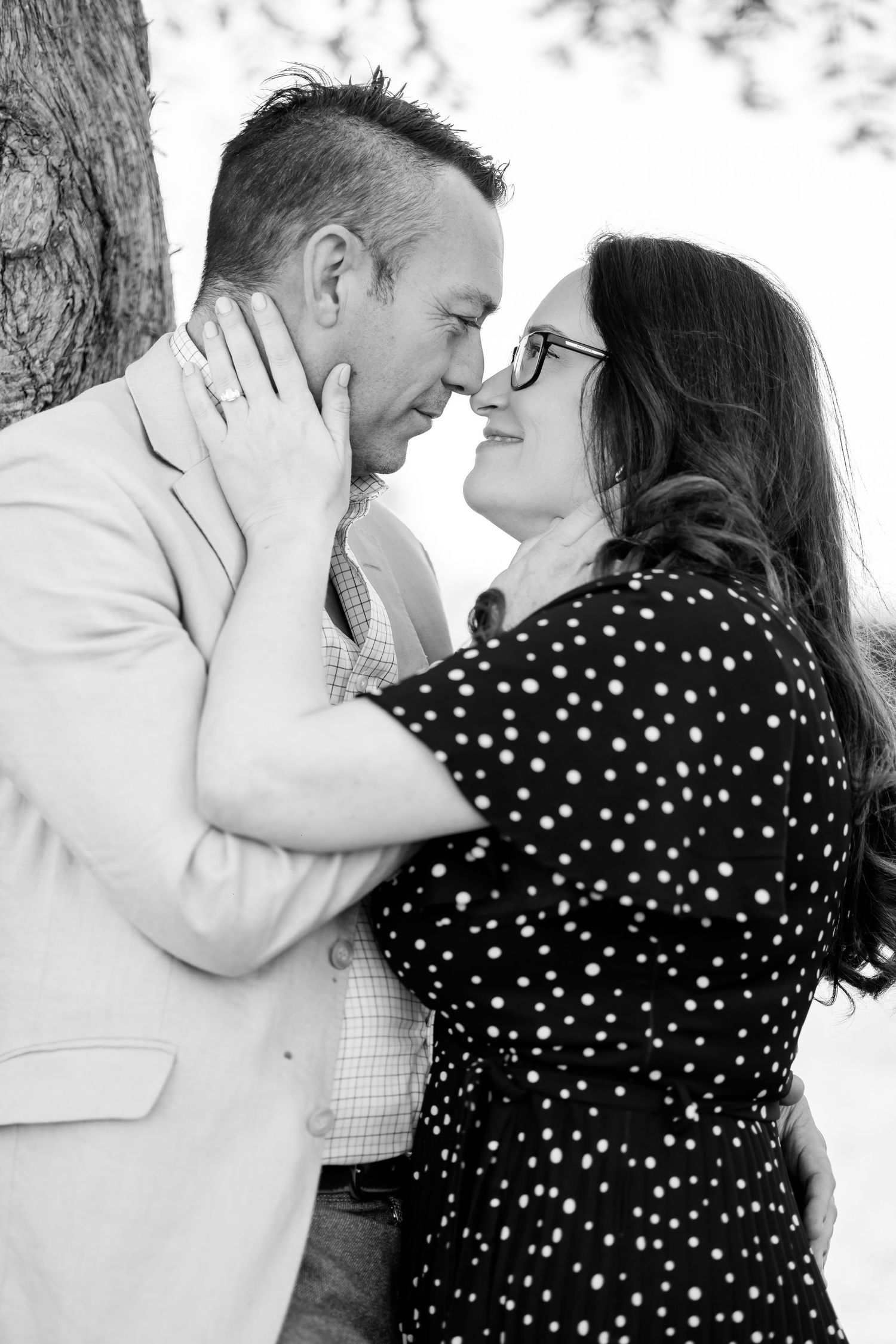 Black and white photo of a couple sharing a romantic moment, leaning in for a kiss near a tree.