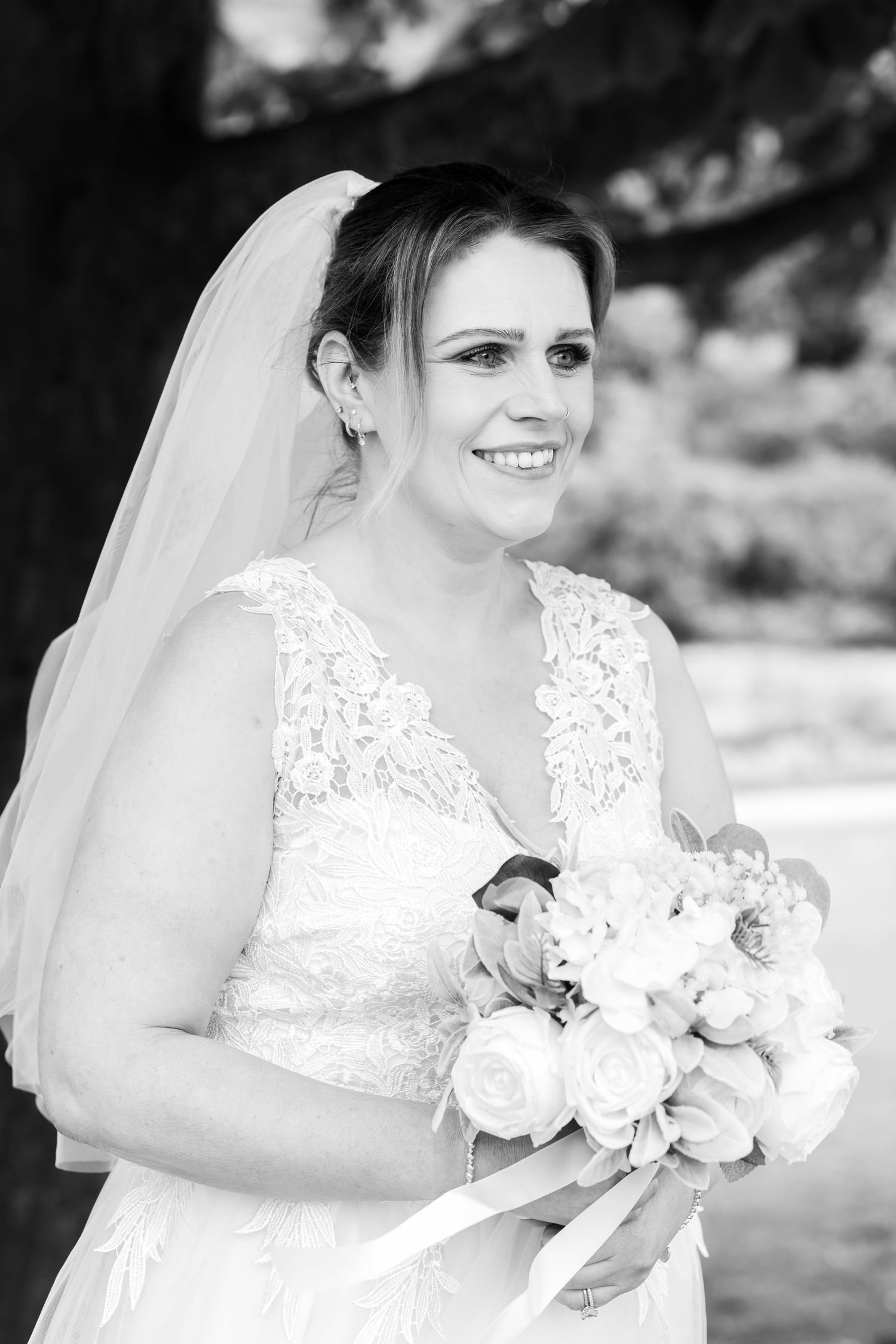 Black and white bridal portrait of smiling bride in lace gown and veil holding white rose bouquet outdoors.