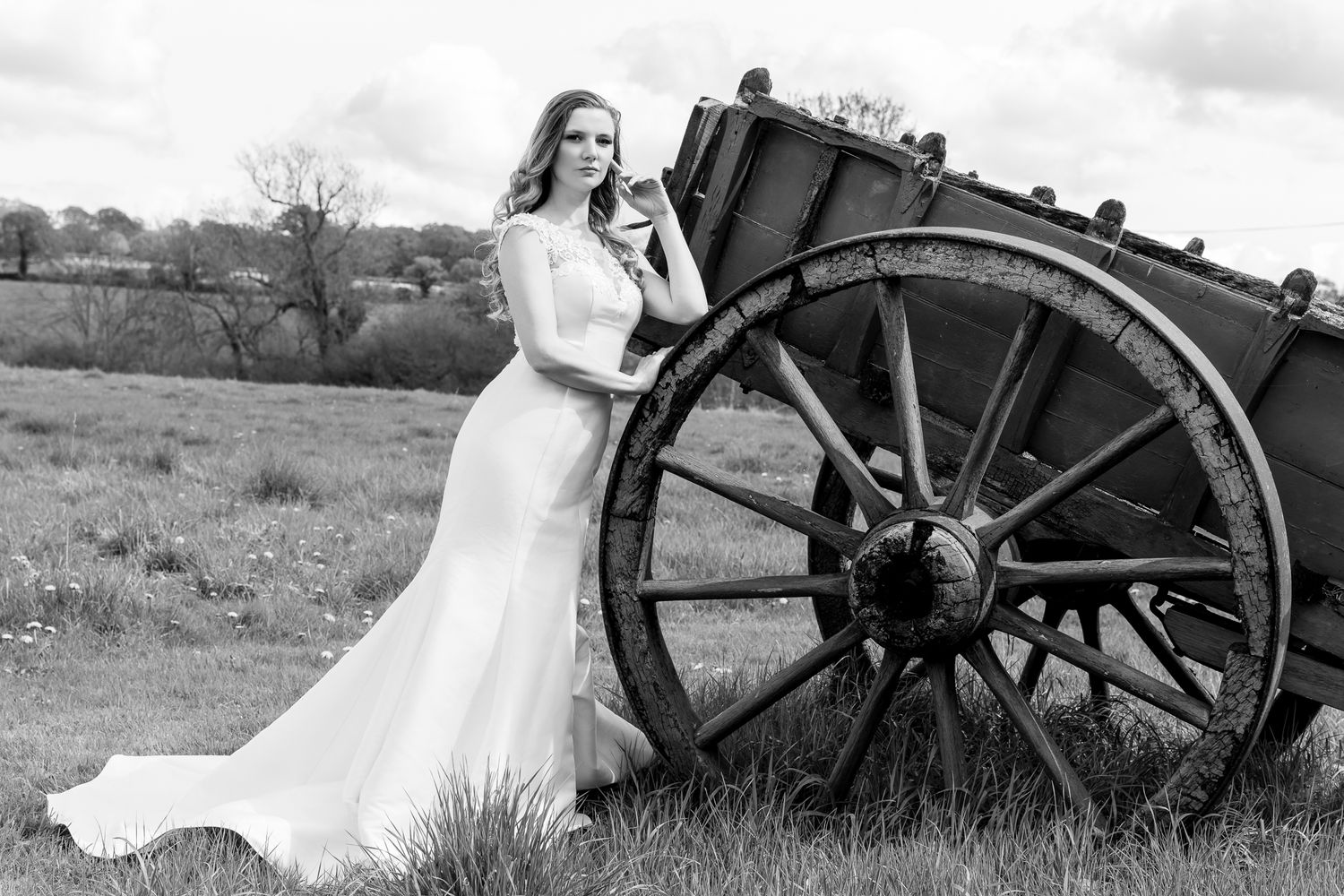 Bride in elegant white gown poses beside a rustic wooden wagon wheel in a black and white outdoor portrait.
