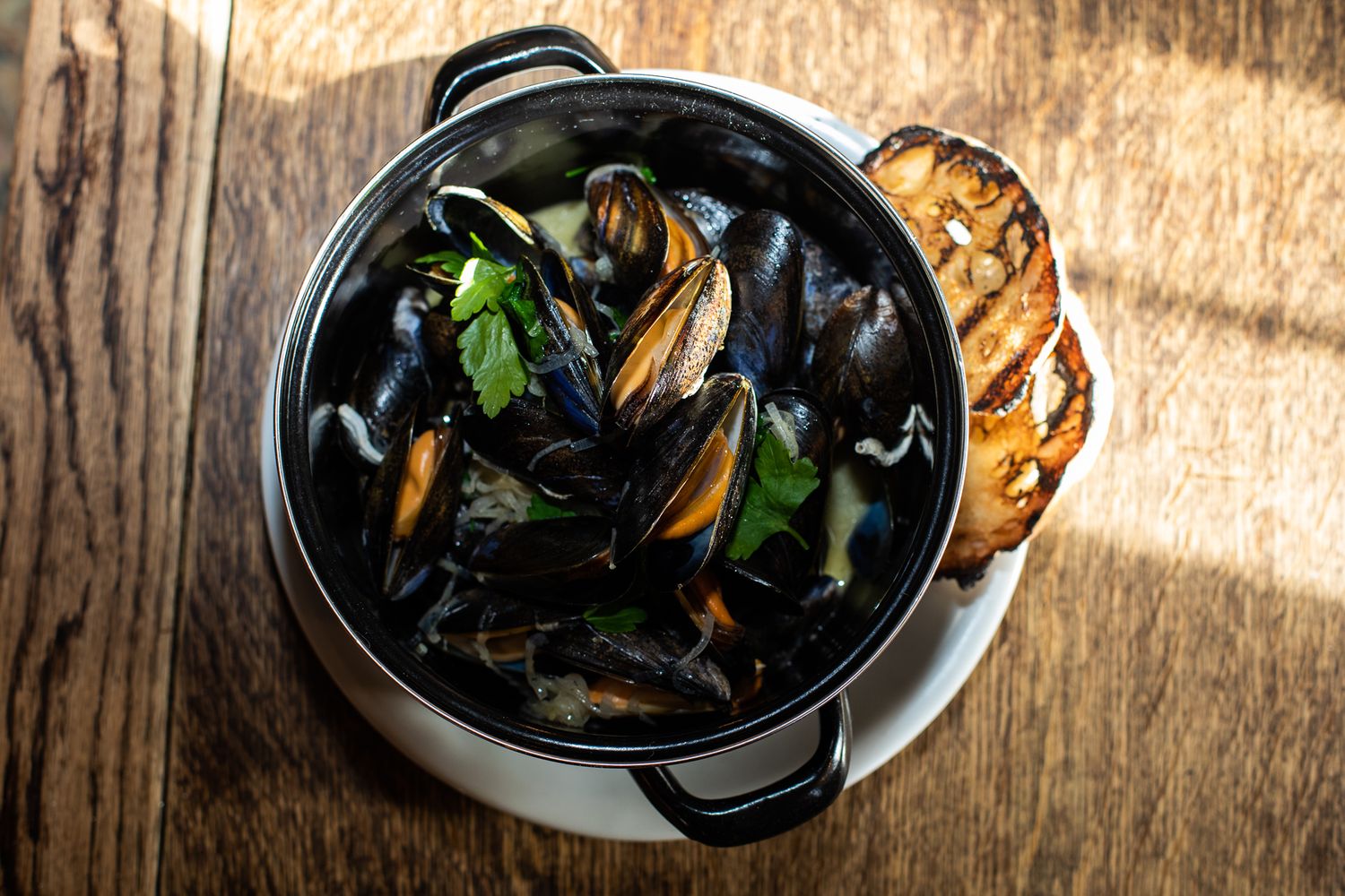 Black pot of steamed mussels with parsley and toasted bread on wooden table.