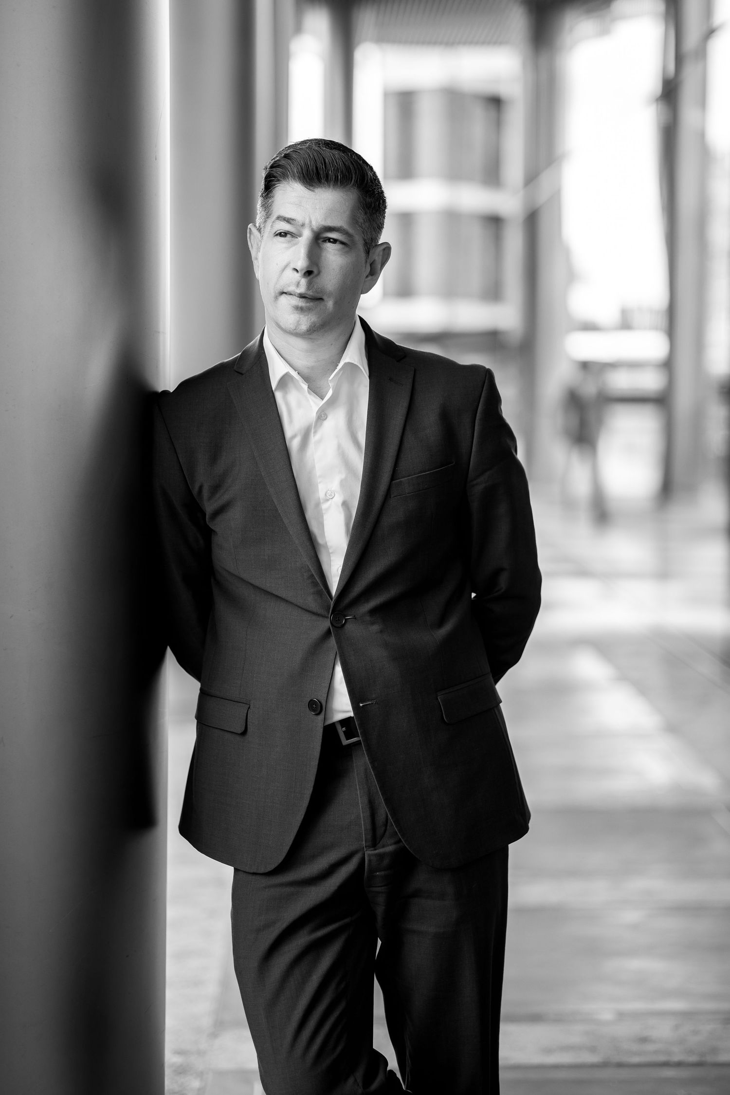 Black and white portrait of a businessman in a dark suit leaning against a column in an urban setting.