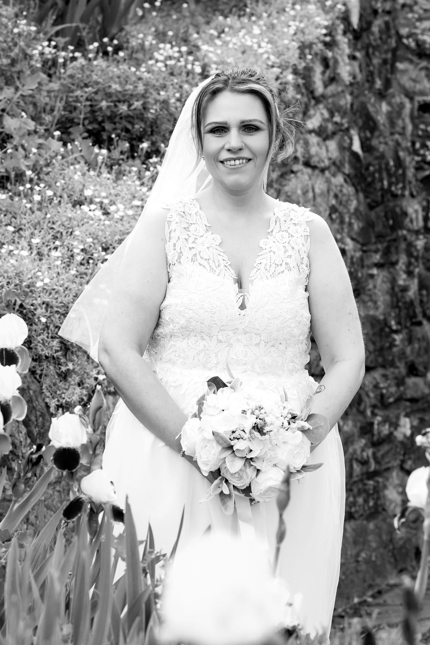 Smiling bride in lace wedding dress and veil holds bouquet of white roses in a lush garden setting.