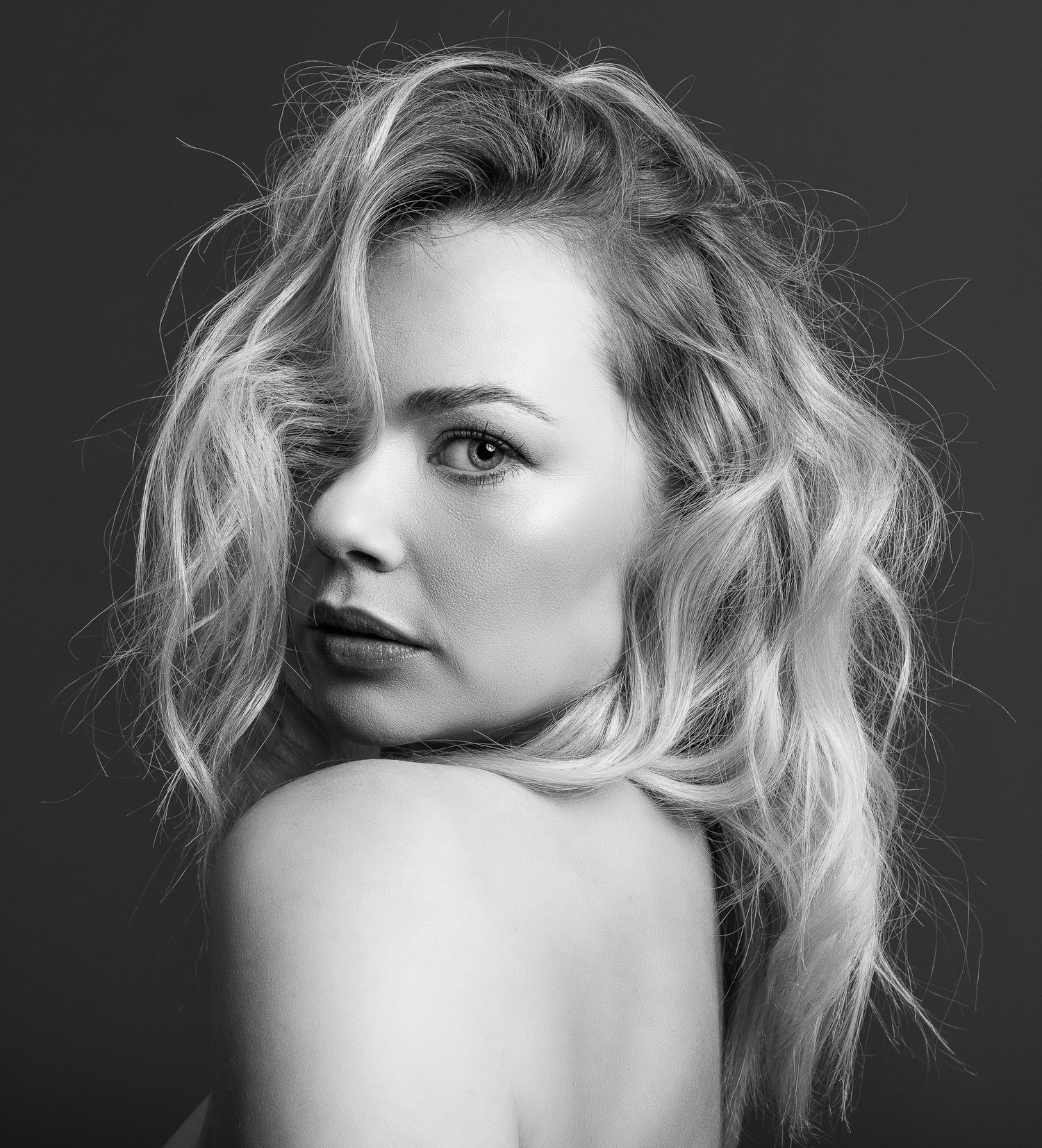 Black and white portrait of a blonde woman with wavy hair looking over her bare shoulder.