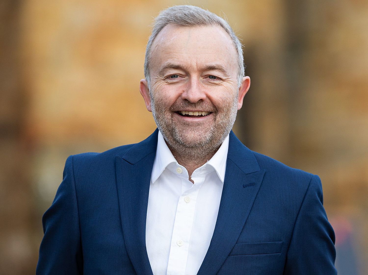 Smiling middle-aged man in a navy blazer and white shirt posing outdoors with autumn foliage background.