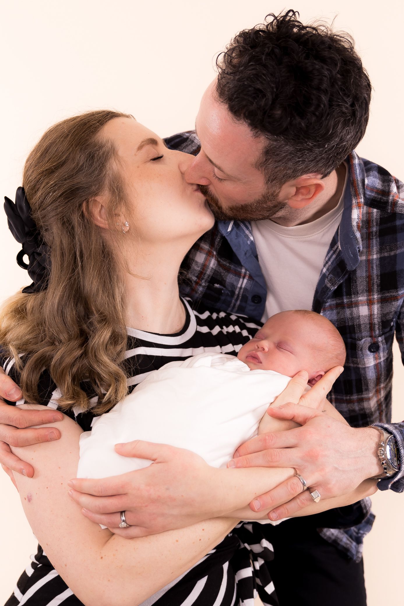 New parents kissing while holding their swaddled newborn baby during a studio portrait session.