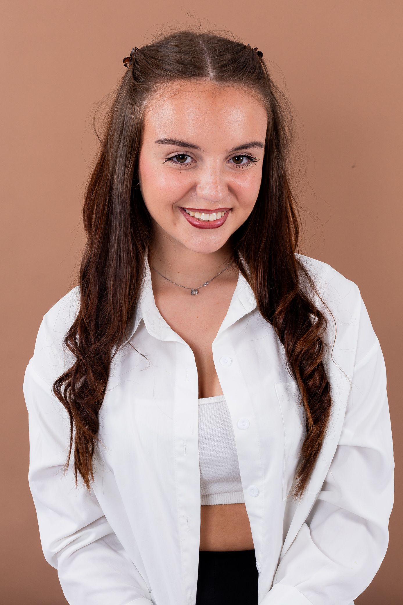 A smiling young woman with long brown hair wears a white shirt and crop top on a tan background.