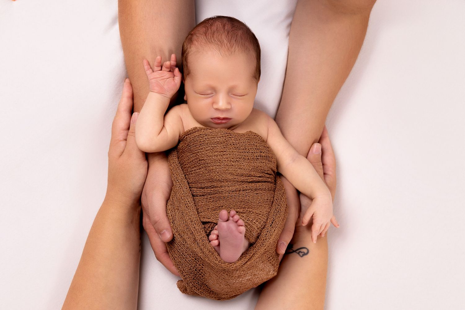 Newborn baby wrapped in brown swaddle cradled in parents hands from above.
