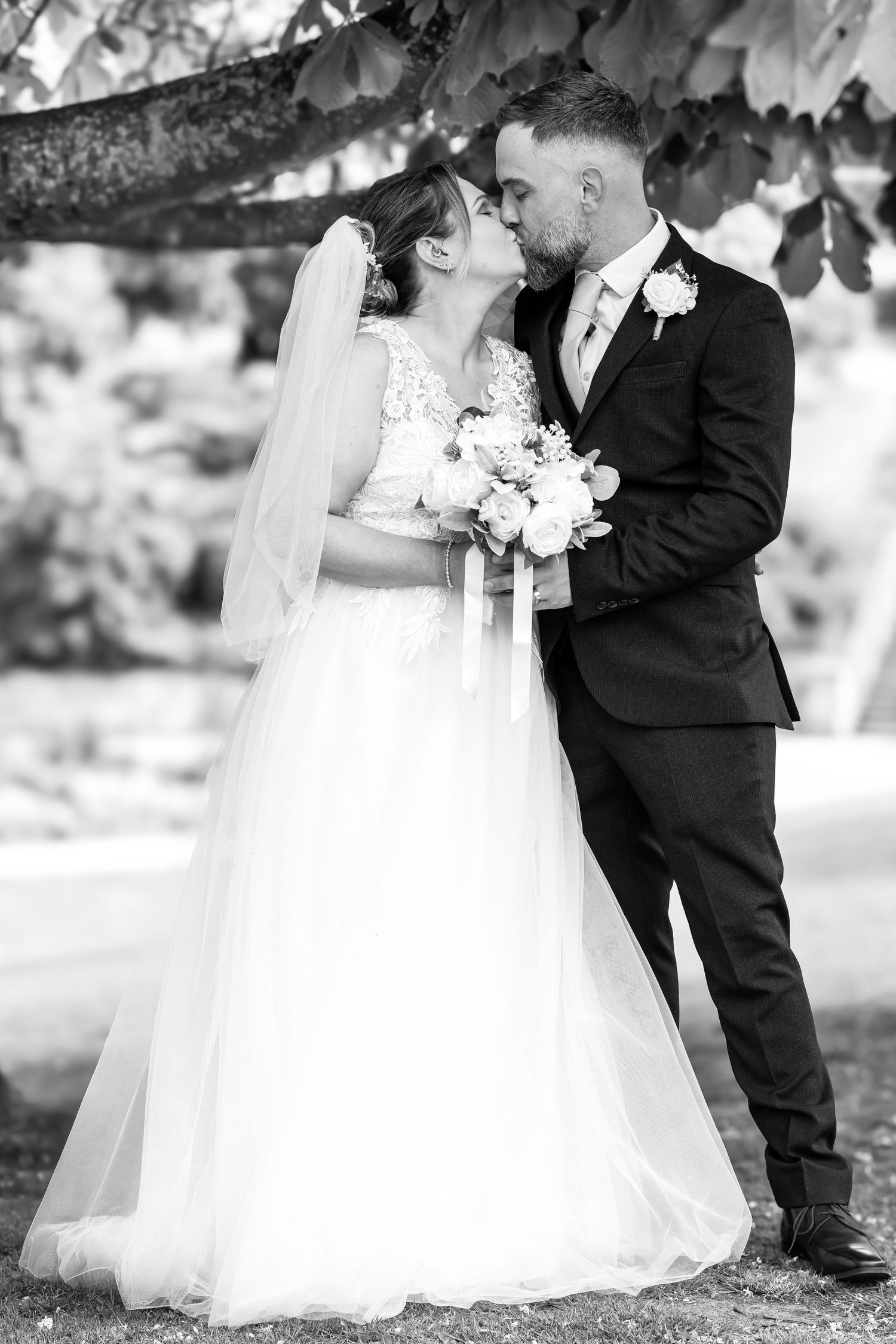 Bride and groom share a romantic kiss outdoors under tree branches in classic black and white wedding portrait.