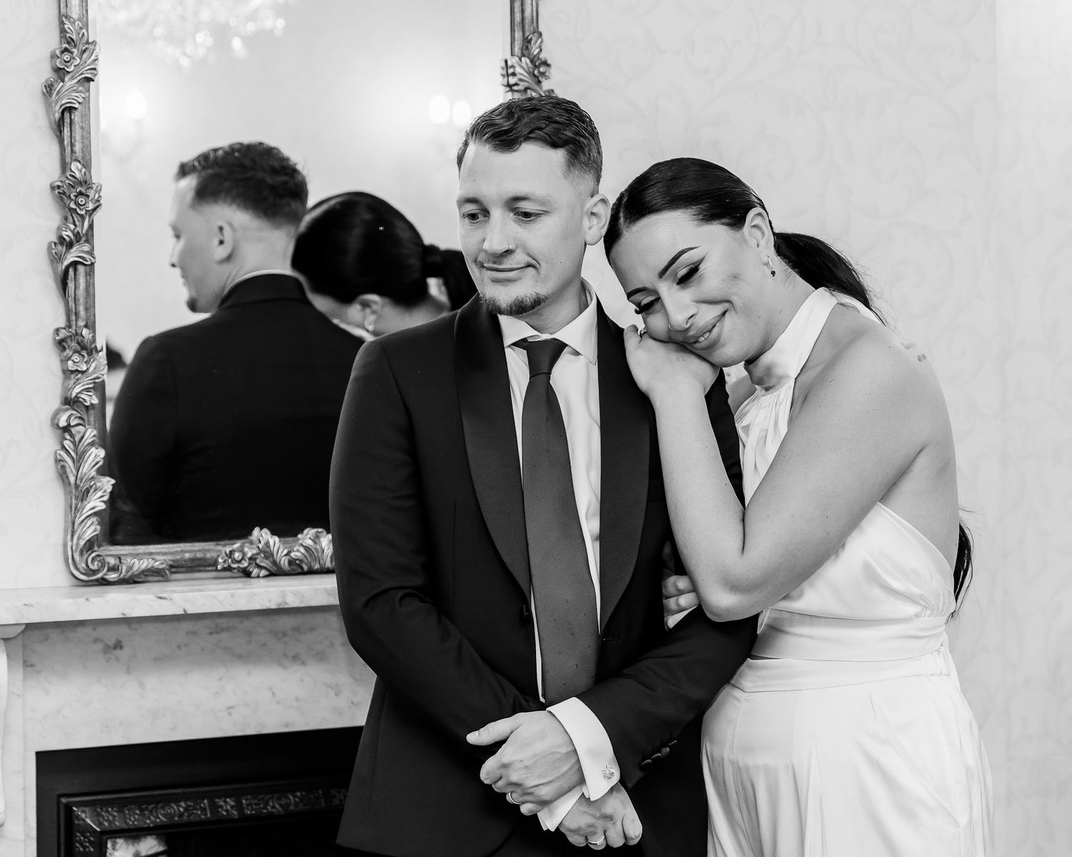 Bride rests head on grooms shoulder in elegant black and white wedding portrait indoors.