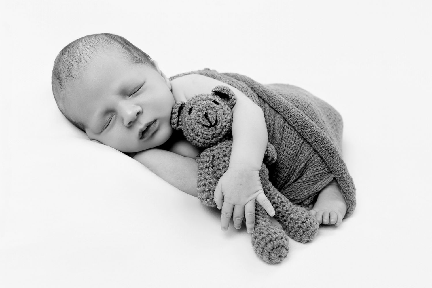 Sleeping newborn baby in knit romper cuddling a small crocheted teddy bear.