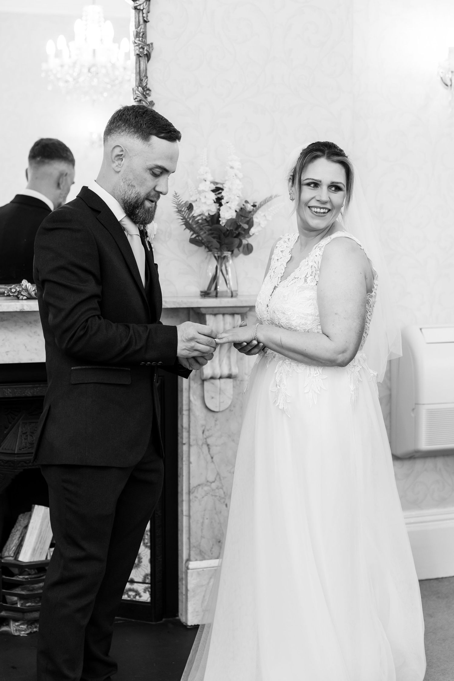 Black and white photo of a groom placing a wedding ring on his smiling bride's finger during their ceremony.