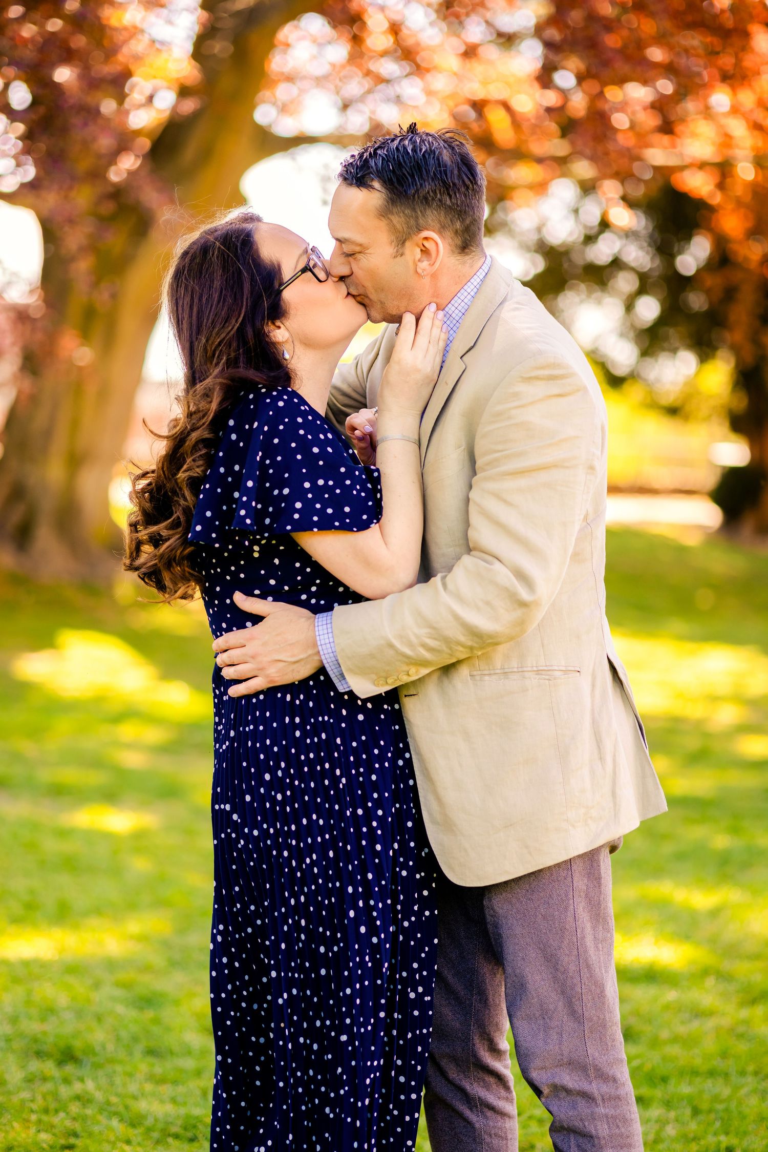 Couple sharing a romantic kiss in a sunlit park, woman in navy polka dot dress, man in beige blazer, autumn trees behind.