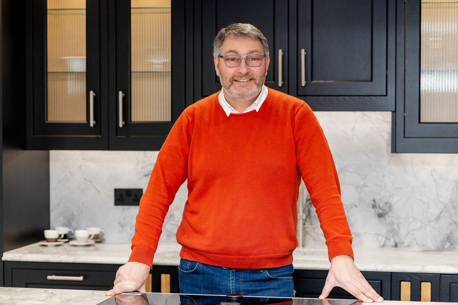 Man in orange sweater smiling in a modern kitchen with dark cabinets and marble countertops.