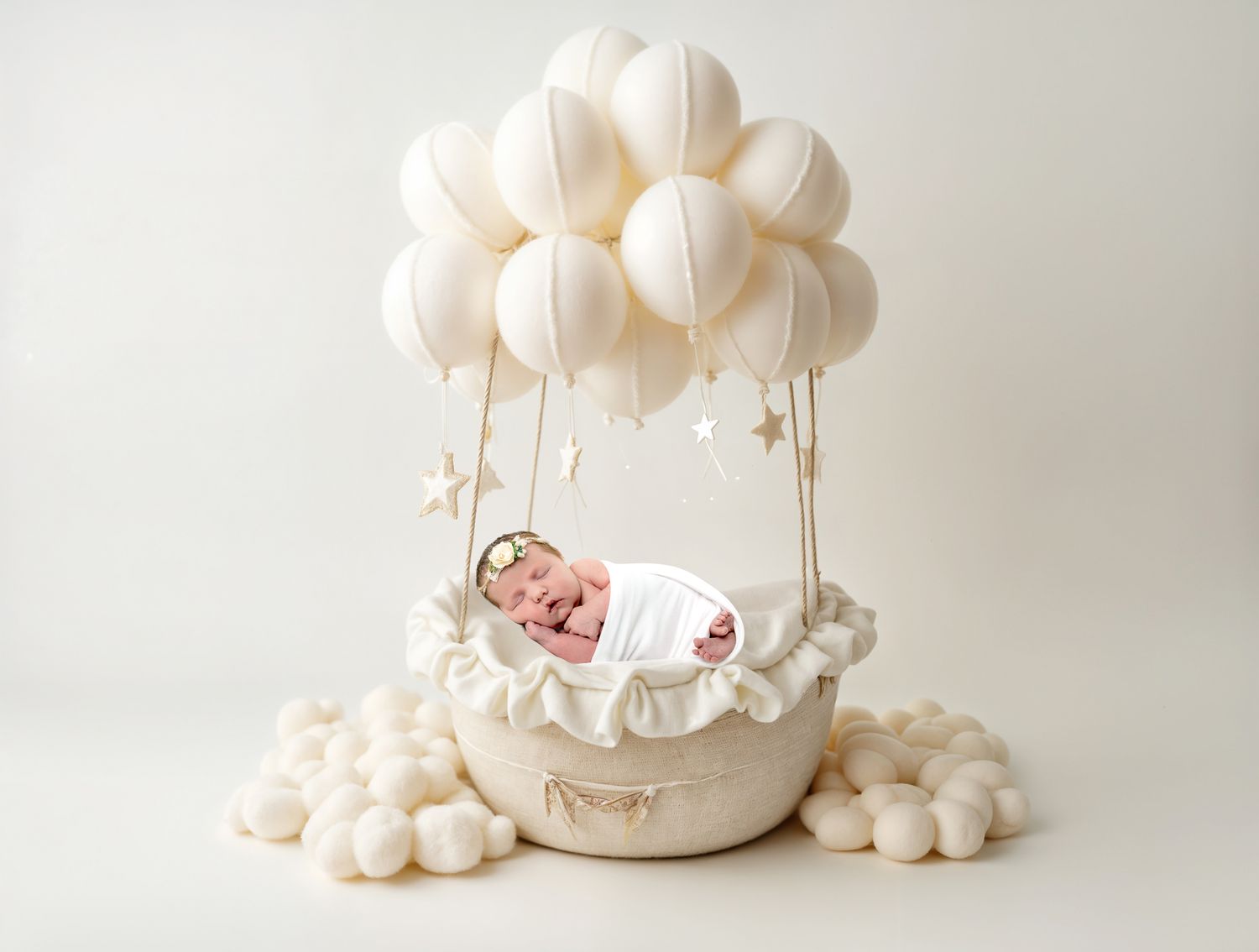 A smiling newborn baby resting in a bowl surrounded by cream balloons and stars in a studio photo.
