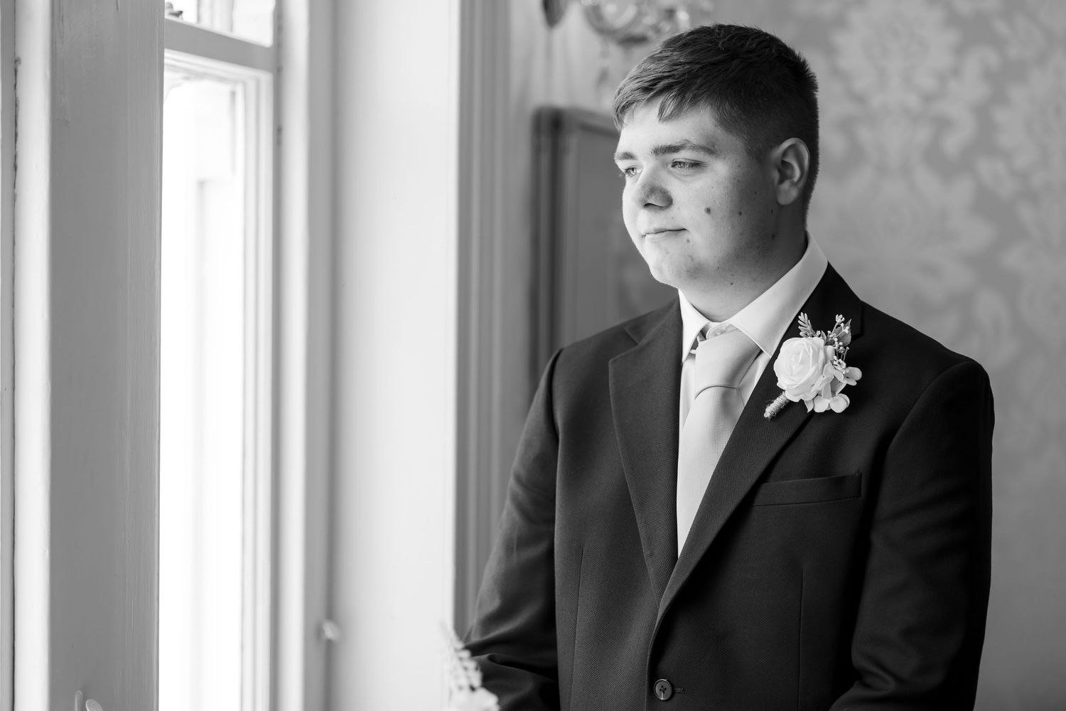 Young groomsman in black suit with floral boutonniere gazes out window in elegant black and white portrait.