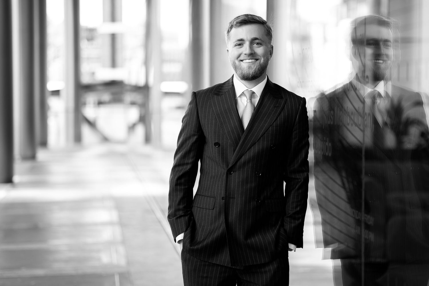 A confident businessman in a pinstripe suit smiles while standing in a modern corporate building corridor.