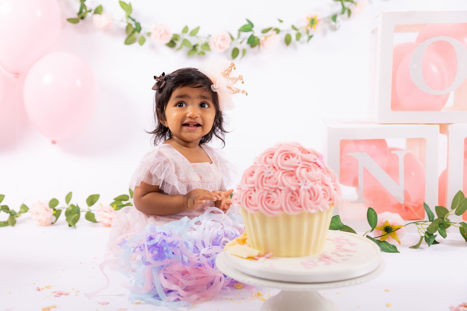 Toddler girl in white dress at cake smash with pink rose cupcake, balloons, and floral blocks on white backdrop.