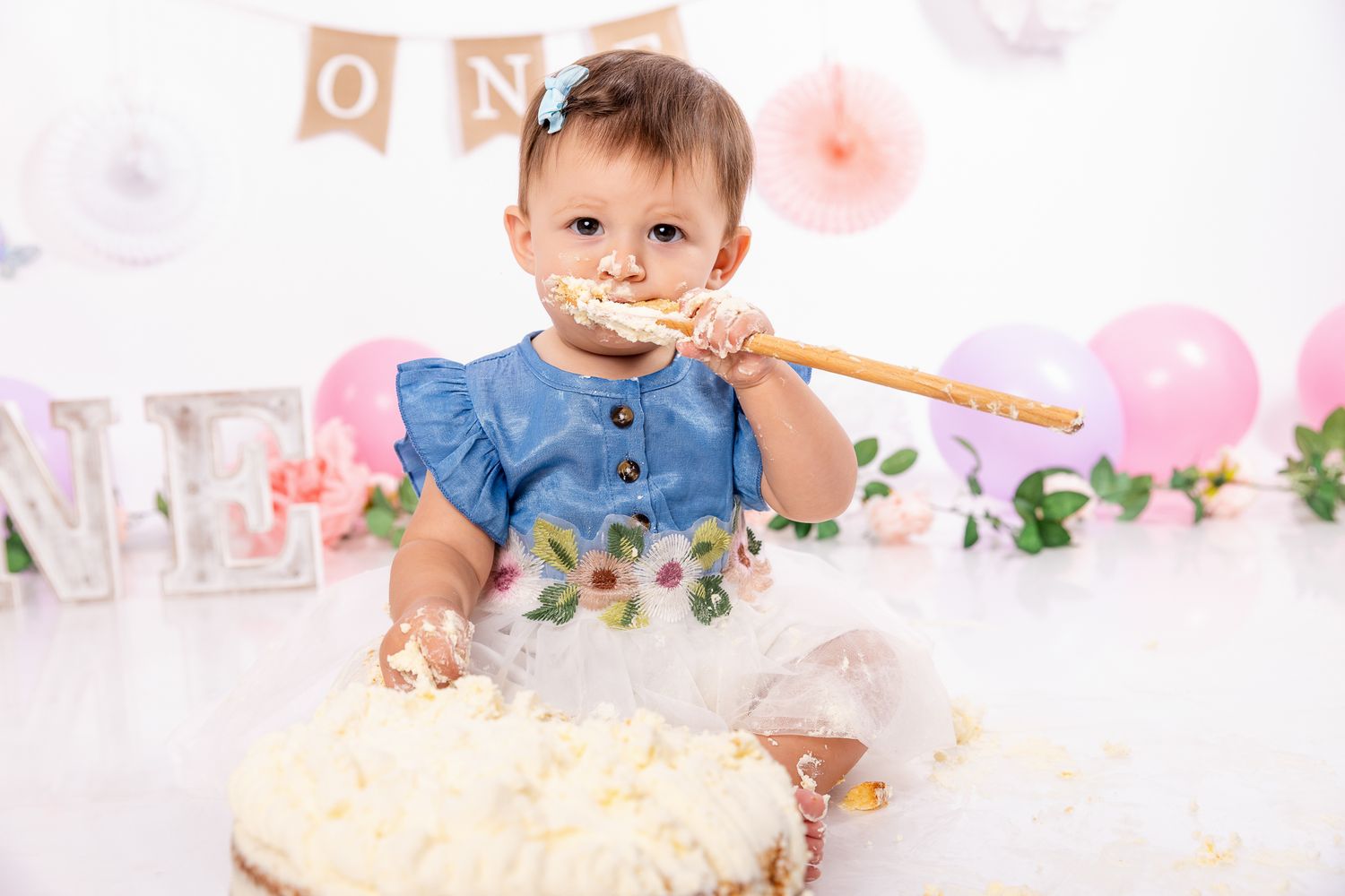 Baby girl in denim dress smashing first birthday cake, sitting among pink balloons and floral decorations.