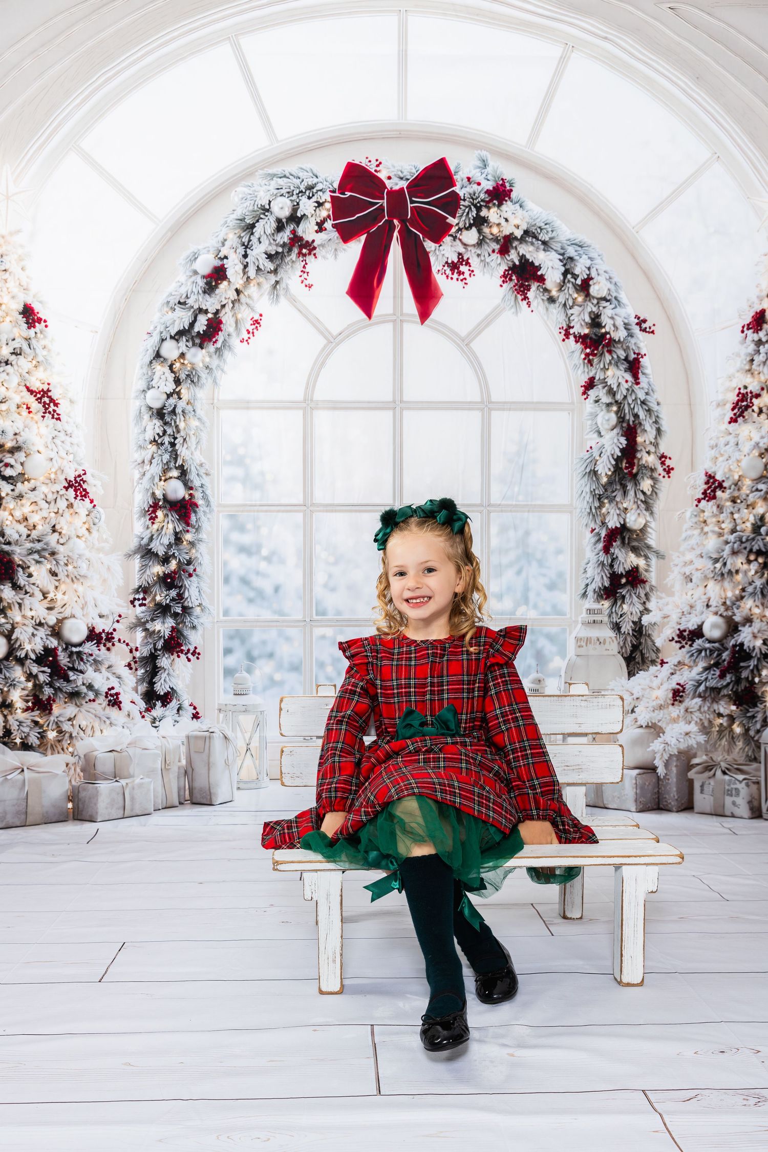 A smiling little girl in a red plaid Christmas dress sits on a white bench surrounded by festive holiday decorations.