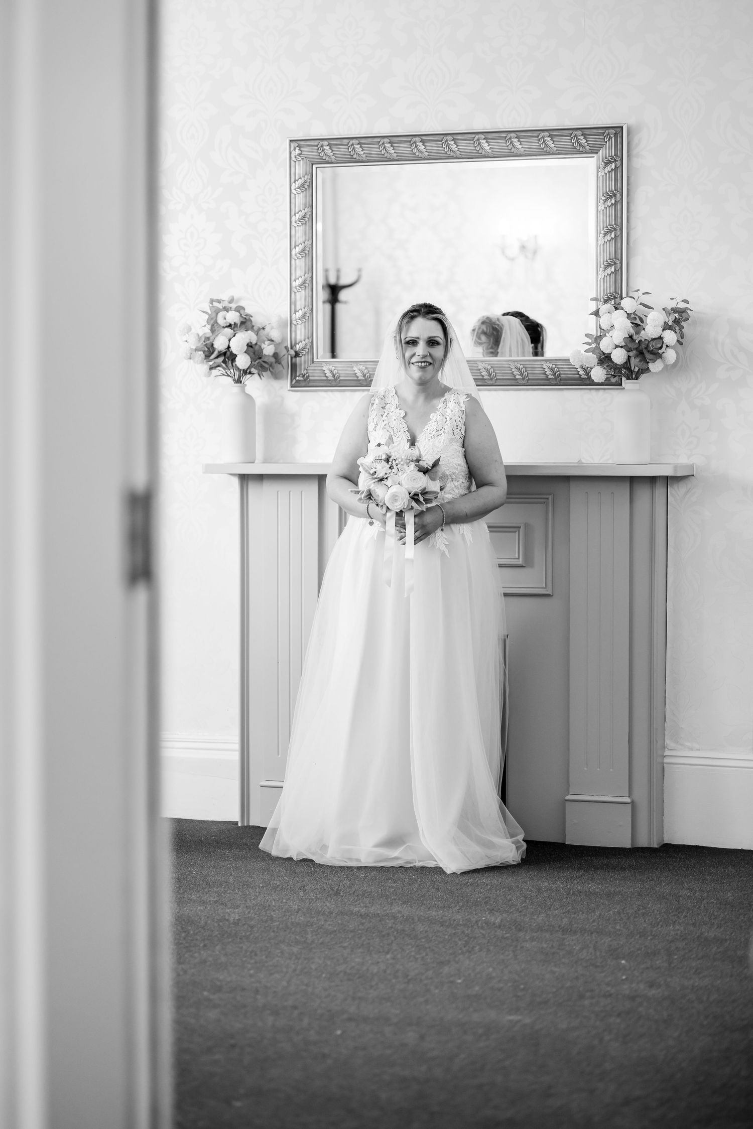 Bride in white gown holding bouquet stands before fireplace with mirror in elegant black and white wedding photo.