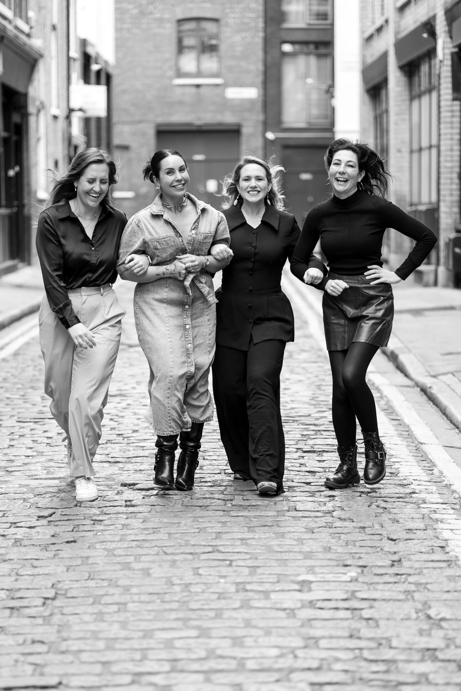 Four women laughing and walking together on a cobblestone street in black and white photo.