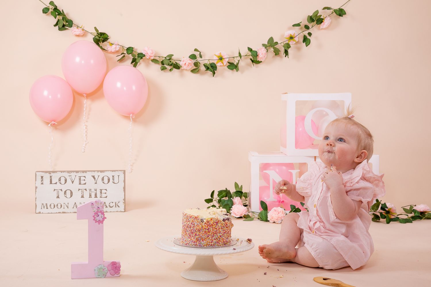 Baby girl first birthday cake smash with pink balloons, floral garland, and decorative blocks on peach background.
