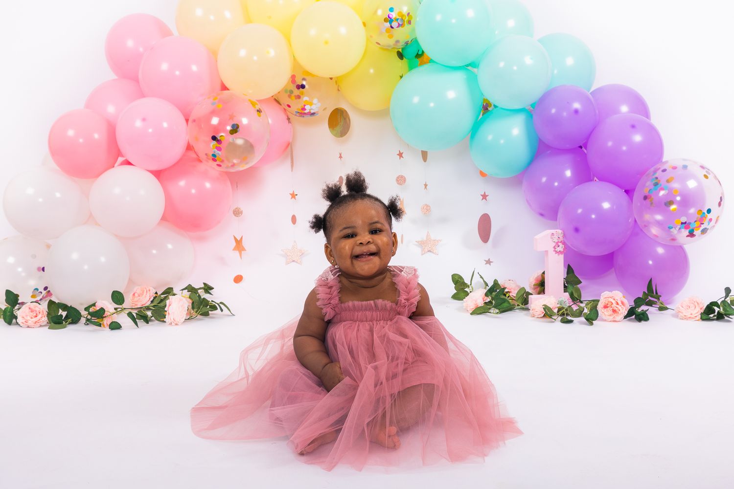 Happy baby girl in pink tutu dress smiling in front of colorful rainbow balloon arch on white background.