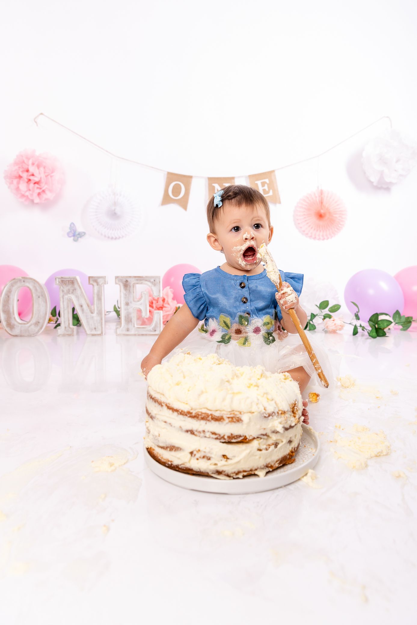 A baby girl in a blue dress smashes her first birthday cake surrounded by ONE decorations and pink balloons.