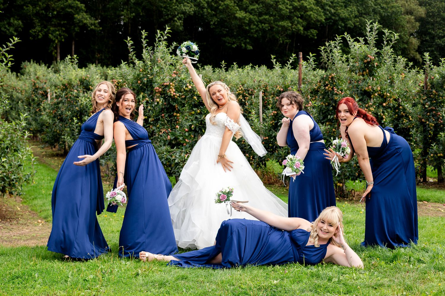 Bride celebrates with five bridesmaids in navy blue dresses in a fun outdoor apple orchard photo.